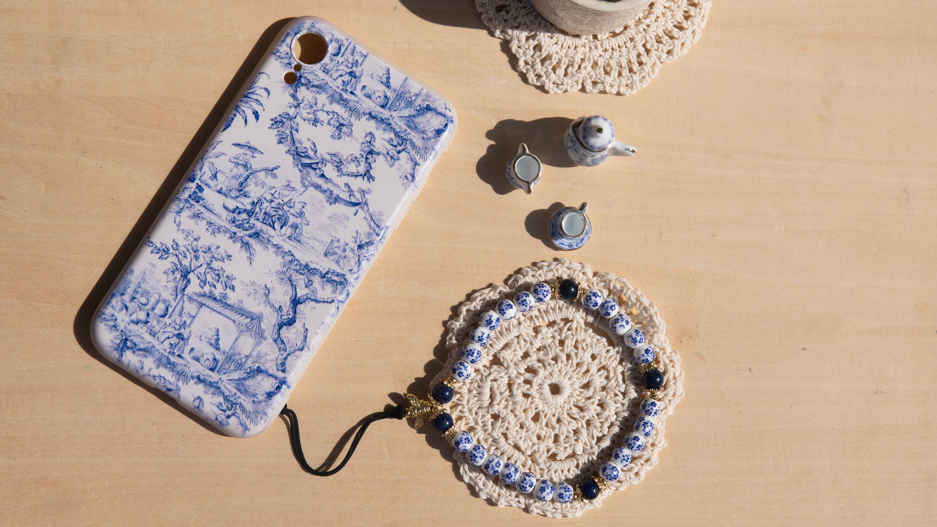 Phone strap attached to a matching phone case - porcelain blue floral pattern on a wooden surface resting on a lace coaster.