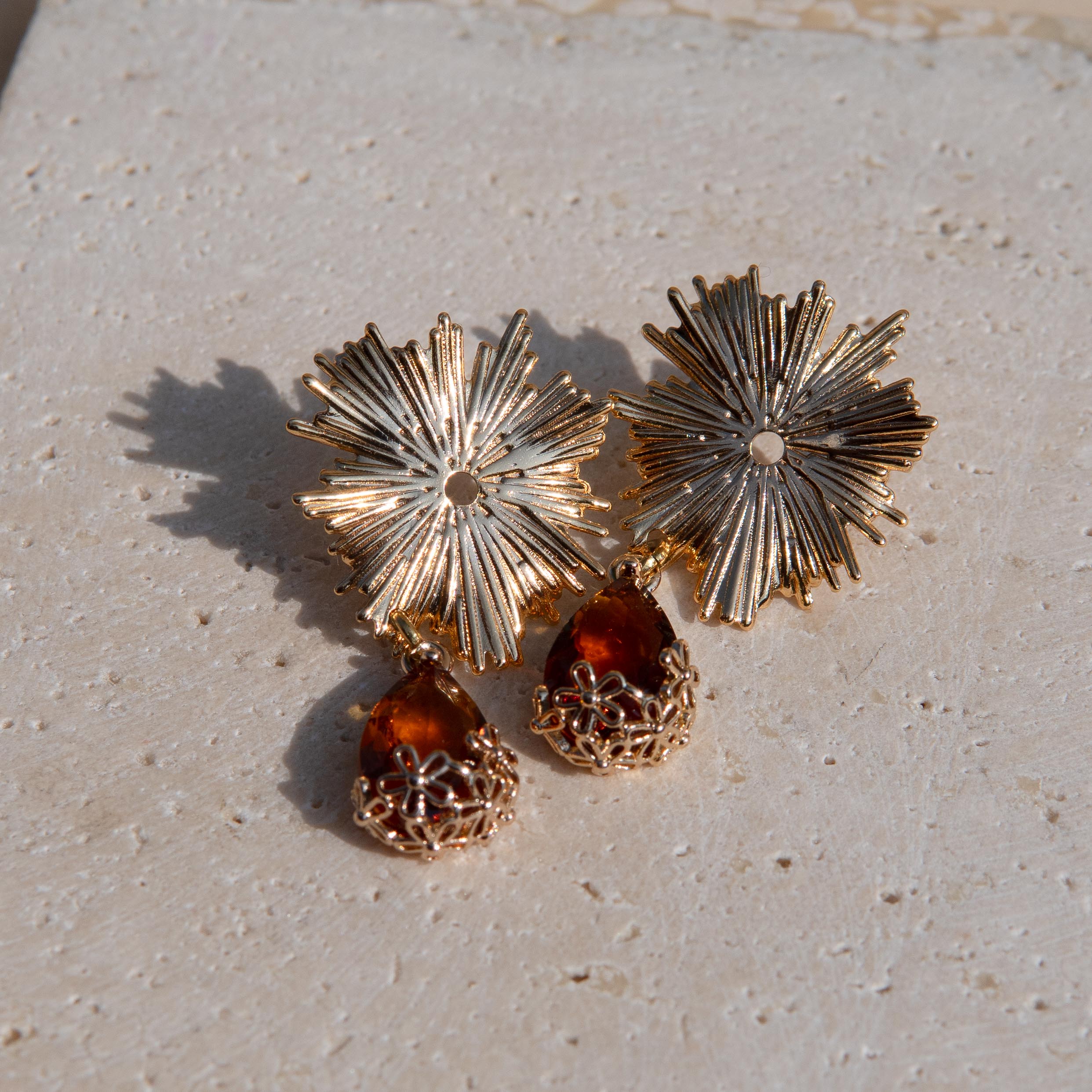 Gold-toned statement earrings with sunburst flower tops and amber teardrop gemstones on a textured beige stone surface.