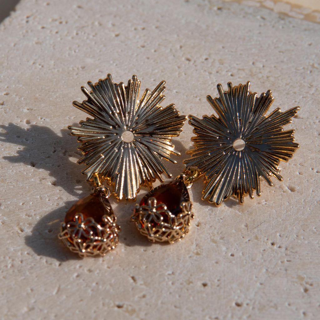 Gold-toned statement earrings with sunburst flower tops and amber teardrop gemstones on a textured beige stone surface.