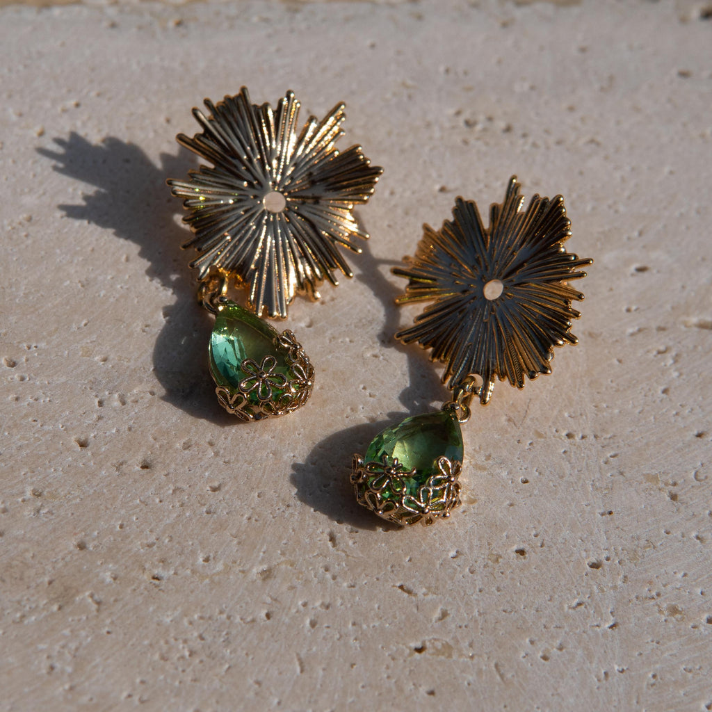 Gold-toned statement earrings with sunburst flower tops and green teardrop gemstones on a textured beige stone surface.