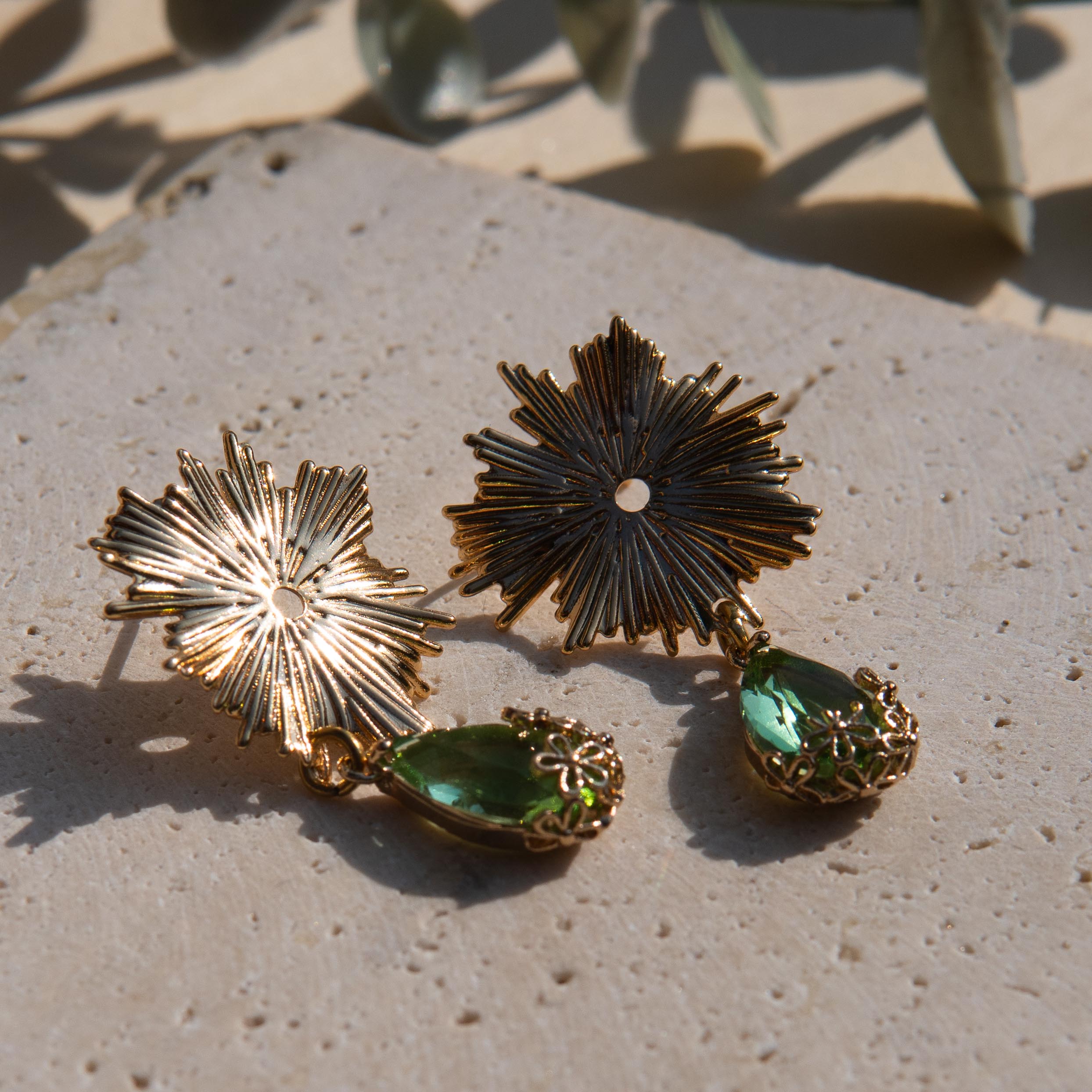 Gold-toned statement earrings with sunburst flower tops and green teardrop gemstones on a textured beige stone surface.