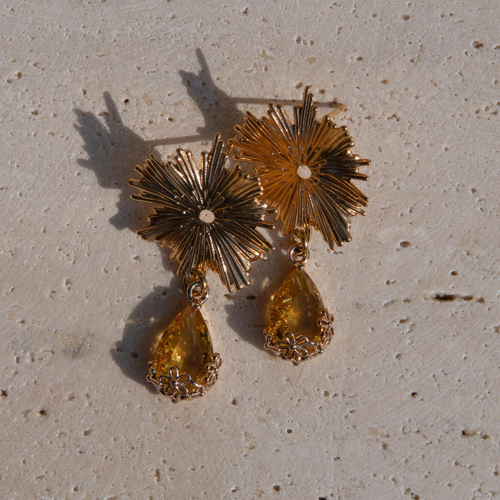 Gold-toned statement earrings with sunburst flower tops and yellow teardrop gemstones on a textured beige stone surface.