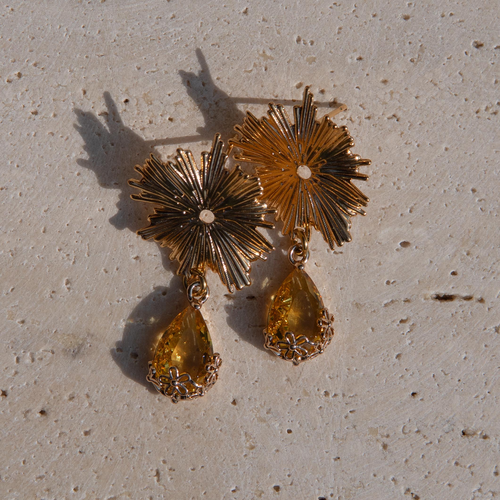 Gold-toned statement earrings with sunburst flower tops and yellow teardrop gemstones on a textured beige stone surface.