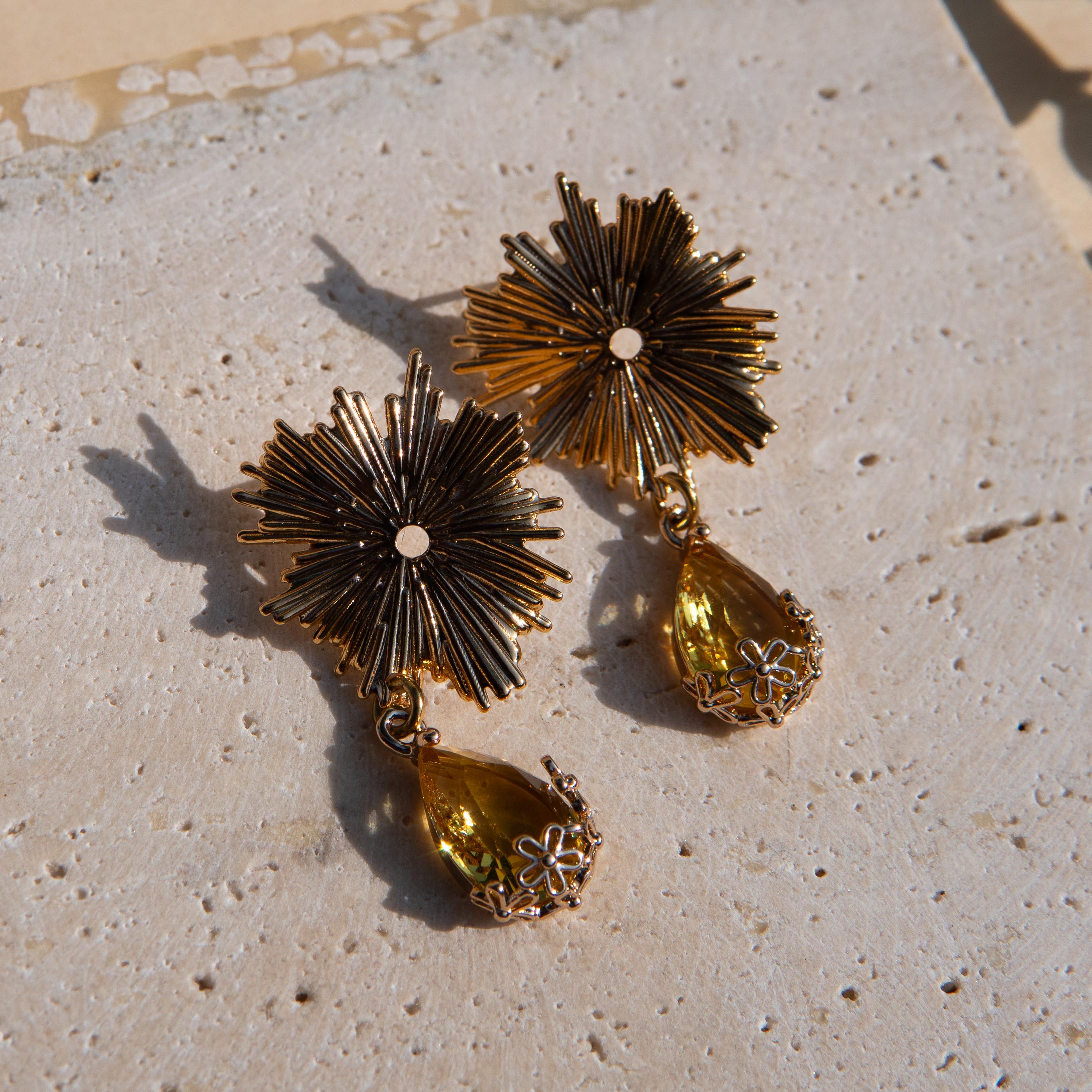 Gold-toned statement earrings with sunburst flower tops and yellow teardrop gemstones on a textured beige stone surface.