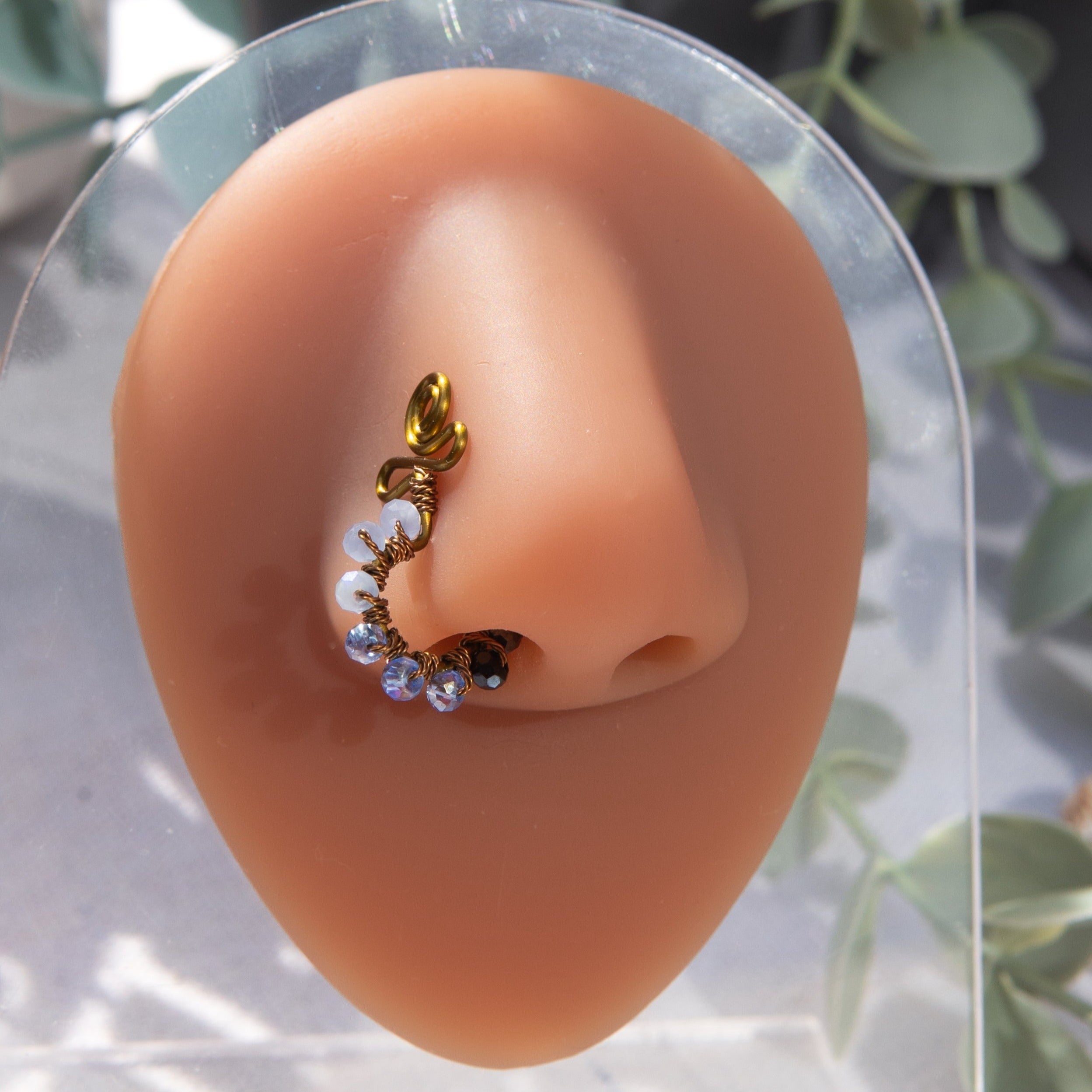 A bronze-toned faux nose ring cuffs the model's nose. Blue transparent seed beads are wire-wrapped around the hoop's circumference. Above the hoop, the wire design forms an Ankh symbol.