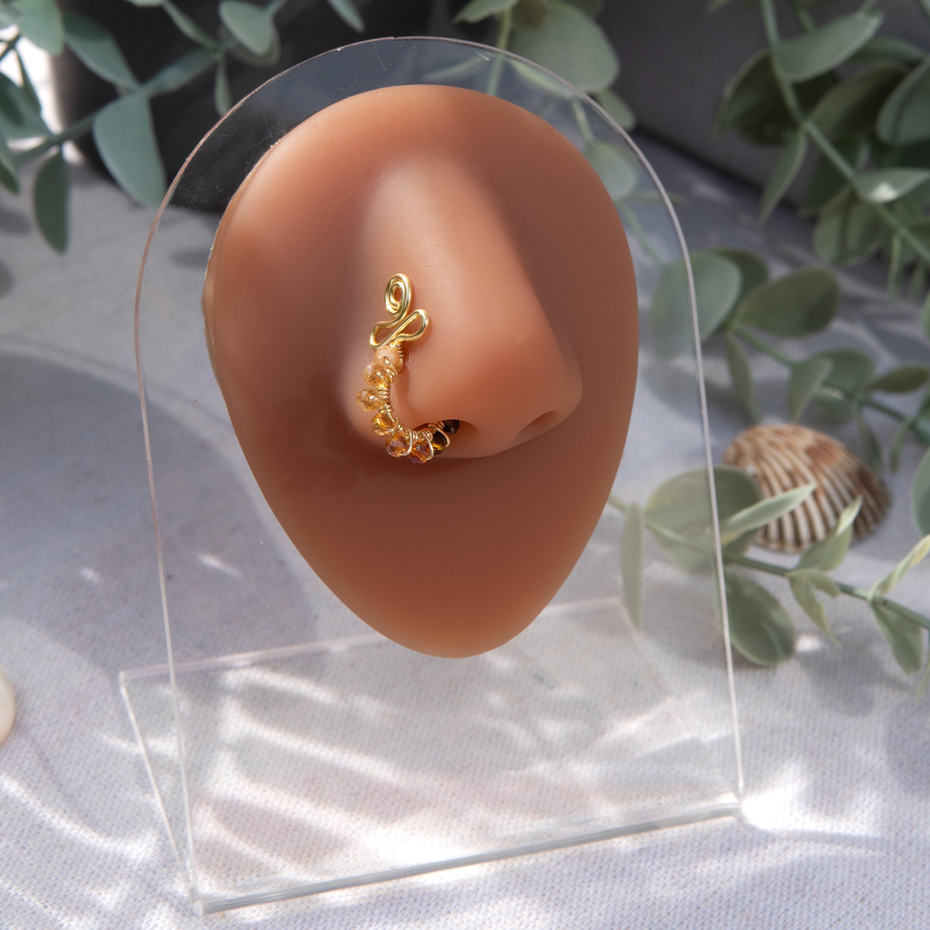 A yellow-gold-toned faux nose ring cuffs the model's nose. Neutral coloured transparent seed beads are wire-wrapped around the hoop's circumference. Above the hoop, the wire design forms an Ankh symbol