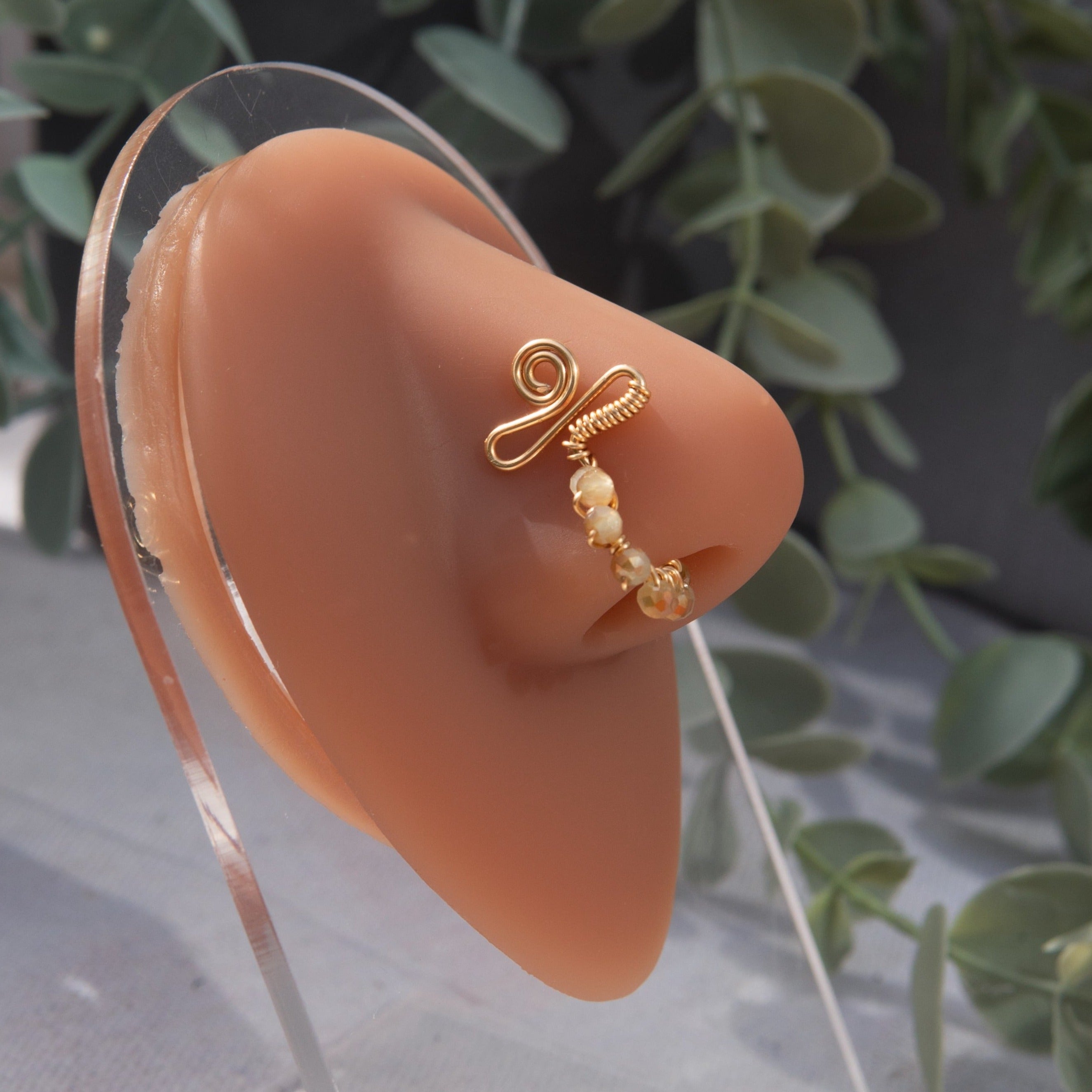 A gold-toned faux nose ring cuffs the model's nose. Transparent seed beads are wire-wrapped around the hoop's circumference. Above the hoop, the wire design forms an Ankh symbol.