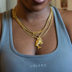 A close-up of a person wearing a layered gold necklace set. The necklace features a gold chain with a 'Blessed' gothic-style pendant, a gold chain with a Queen Nefertiti pendant, and another chain with a  Queen Cleopatra pendant.
