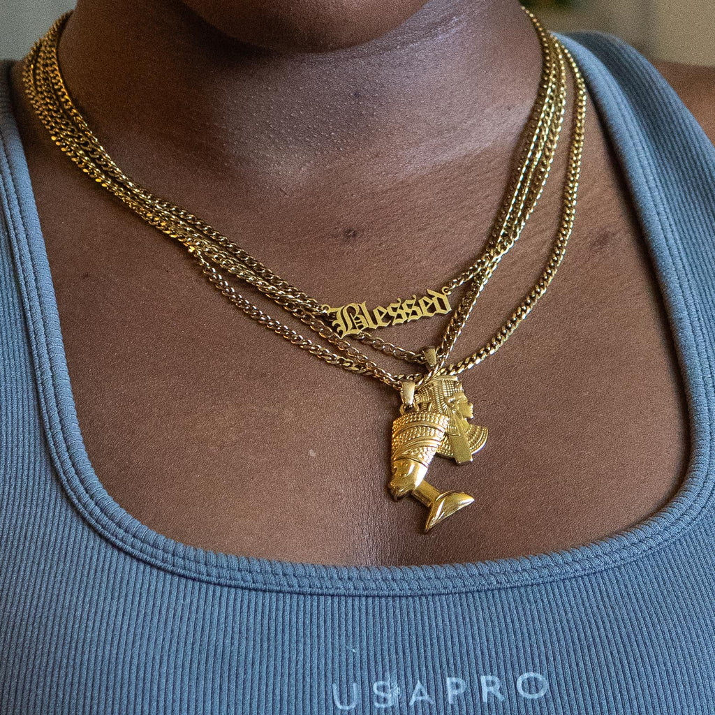A close-up of a person wearing a layered gold necklace set. The necklace features a gold chain with a 'Blessed' gothic-style pendant, a gold chain with a Queen Nefertiti pendant, and another chain with a  Queen Cleopatra pendant.