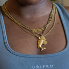 A close-up of a person wearing a layered gold necklace set. The necklace features a gold chain with a 'Blessed' gothic-style pendant, a gold chain with a Queen Nefertiti pendant, and another chain with a  Queen Cleopatra pendant.