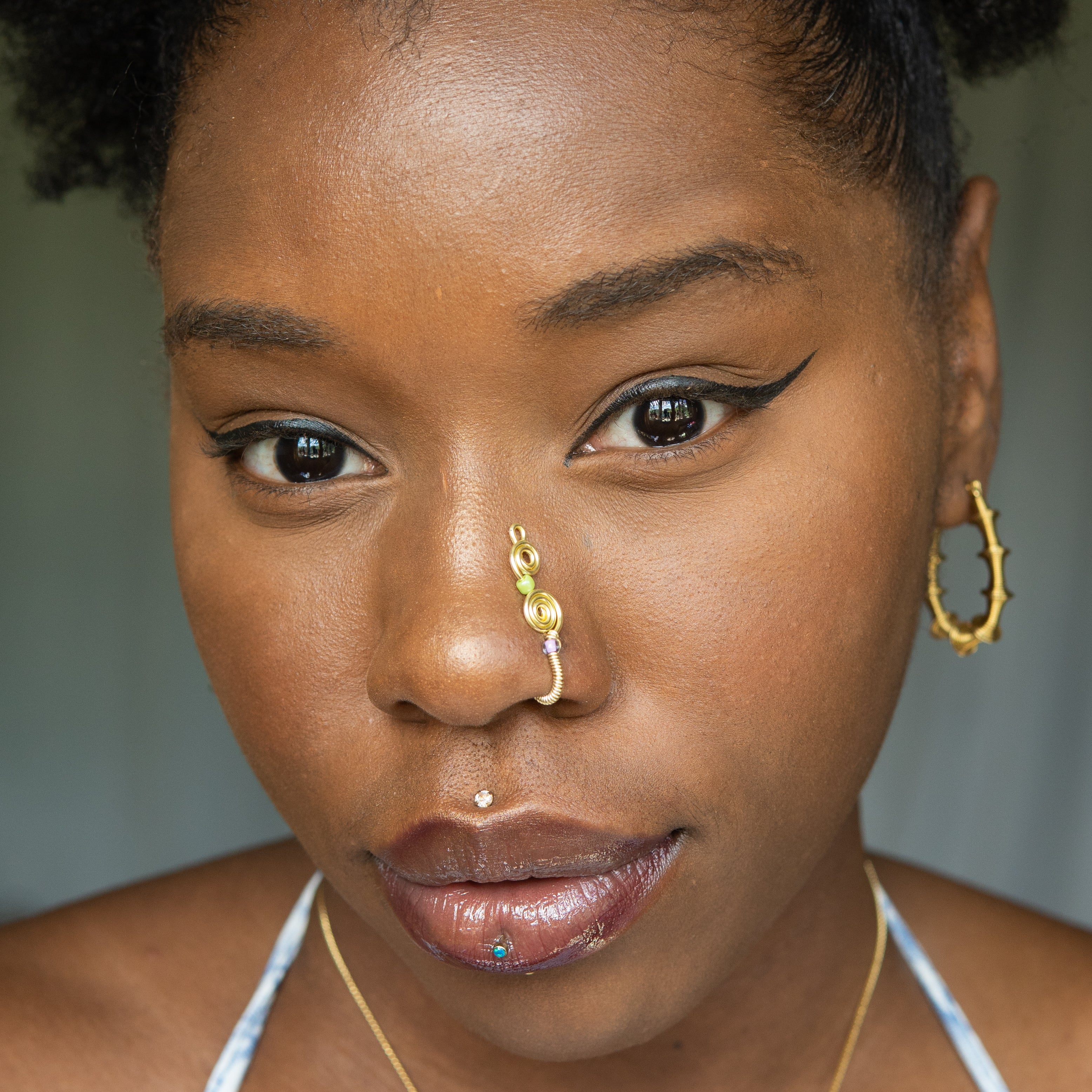 A light gold-toned, tightly spiralled nose cuff rests on the model's nose. Two seed beads (green and lilac) are interwoven into the design as the 3-tier circular spiral gradually decreases in size.
