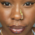 A light gold-toned, tightly spiralled nose cuff rests on the model's nose. Two seed beads (green and lilac) are interwoven into the design as the 3-tier circular spiral gradually decreases in size.