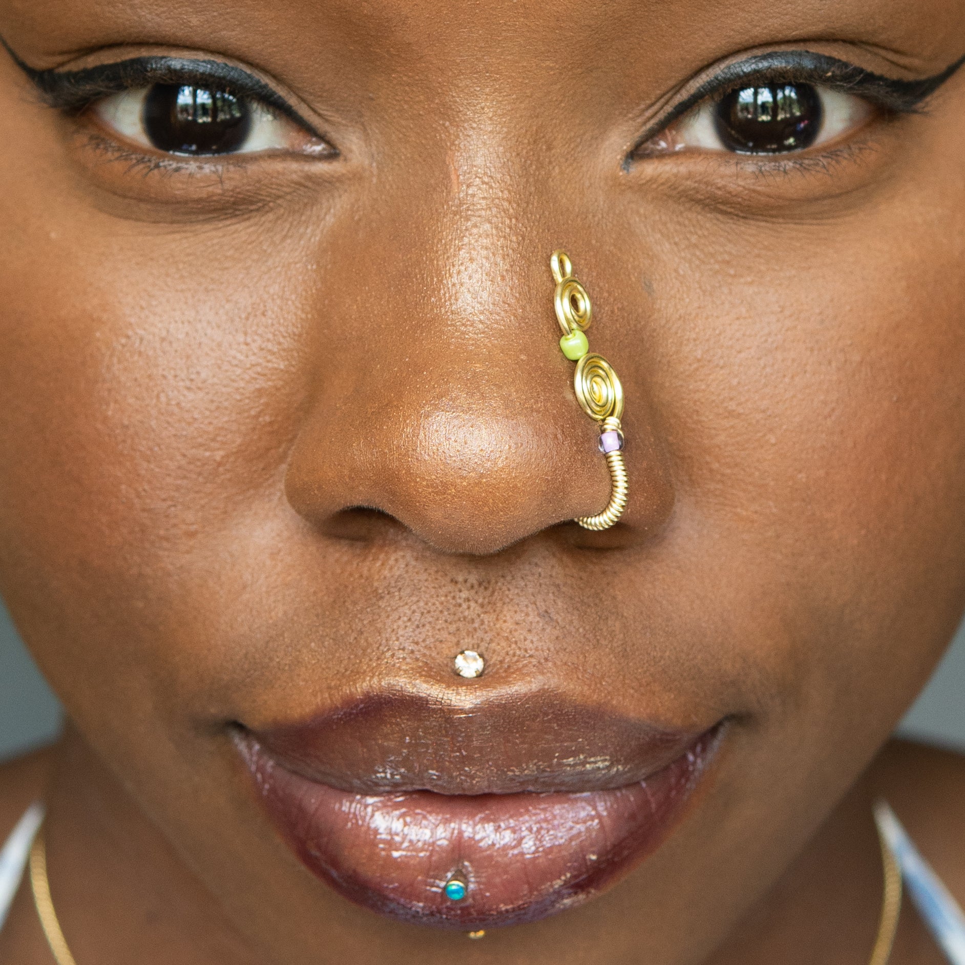 A light gold-toned, tightly spiralled nose cuff rests on the model's nose. Two seed beads (green and lilac) are interwoven into the design as the 3-tier circular spiral gradually decreases in size.