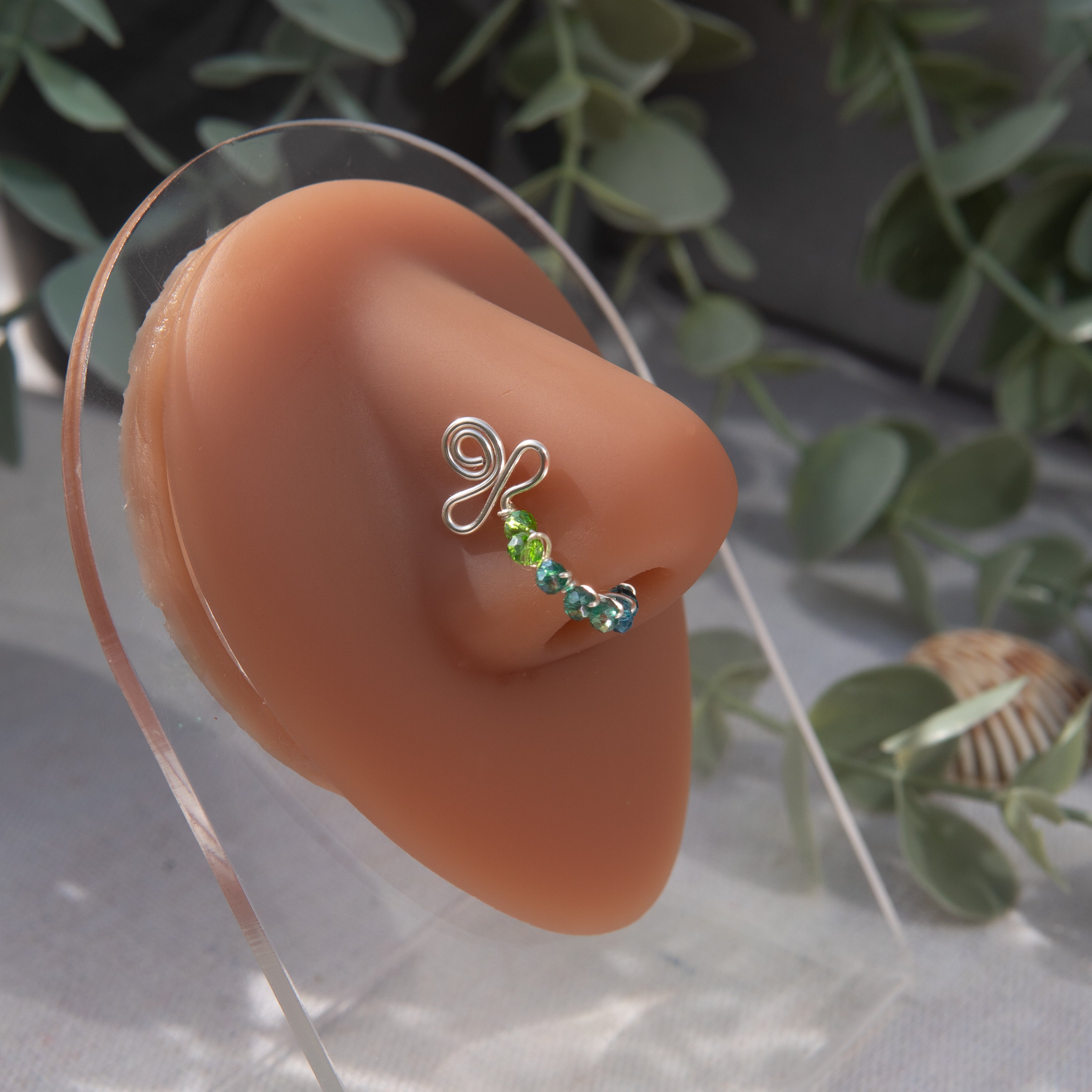 A silver-toned faux nose ring cuffs the model's nose. Transparent seed beads are wire-wrapped around the hoop's circumference. Above the hoop, the wire design forms an Ankh symbol. 
