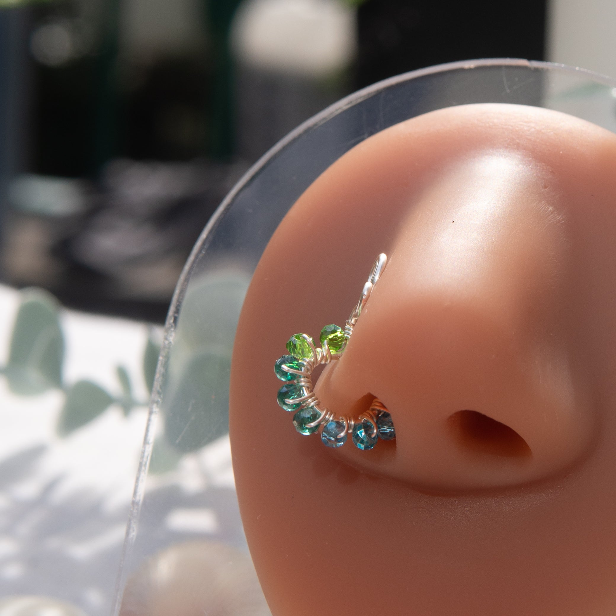 A silver-toned faux nose ring cuffs the model's nose. Transparent seed beads are wire-wrapped around the hoop's circumference. Above the hoop, the wire design forms an Ankh symbol. 
