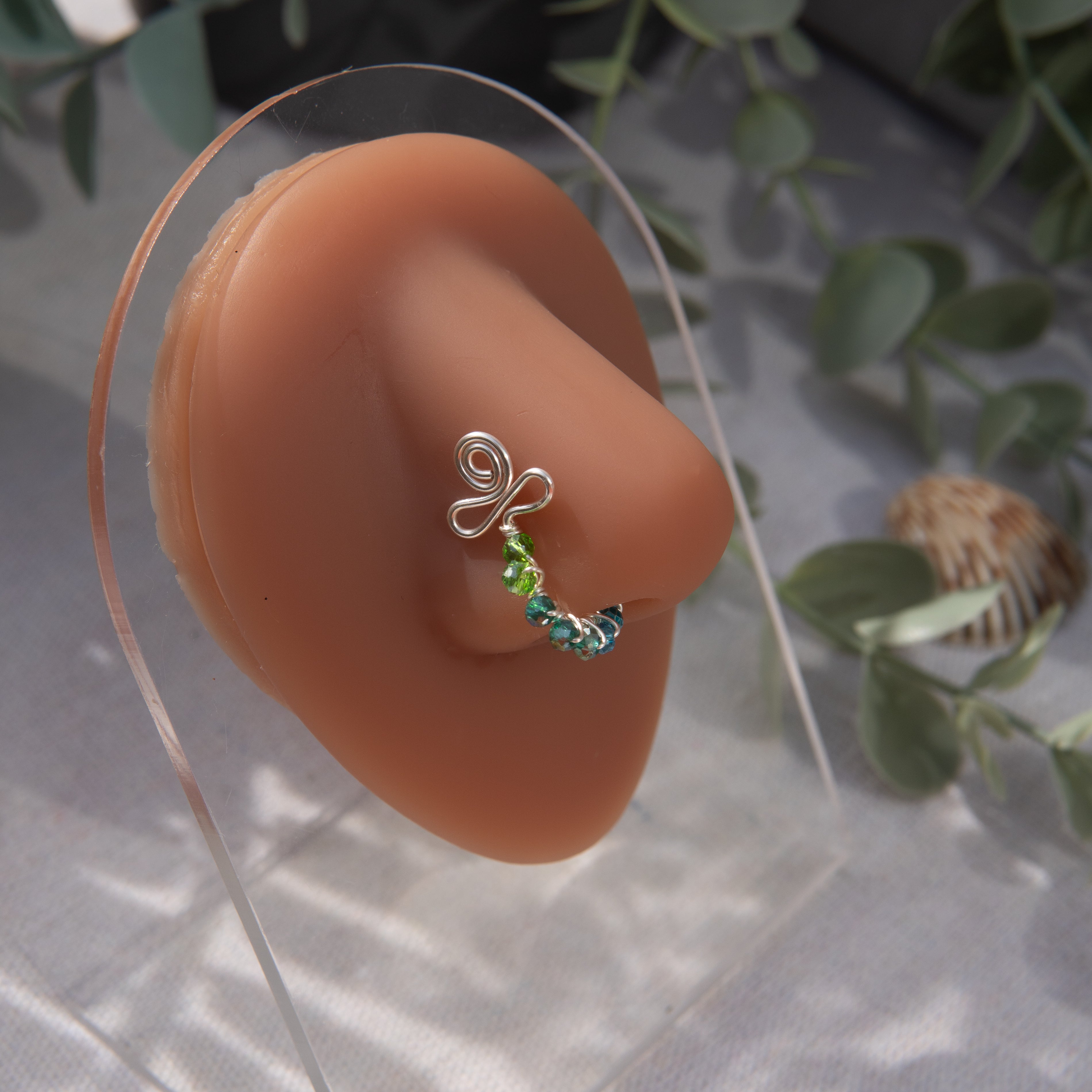 A silver-toned faux nose ring cuffs the model's nose. Transparent seed beads are wire-wrapped around the hoop's circumference. Above the hoop, the wire design forms an Ankh symbol. 
