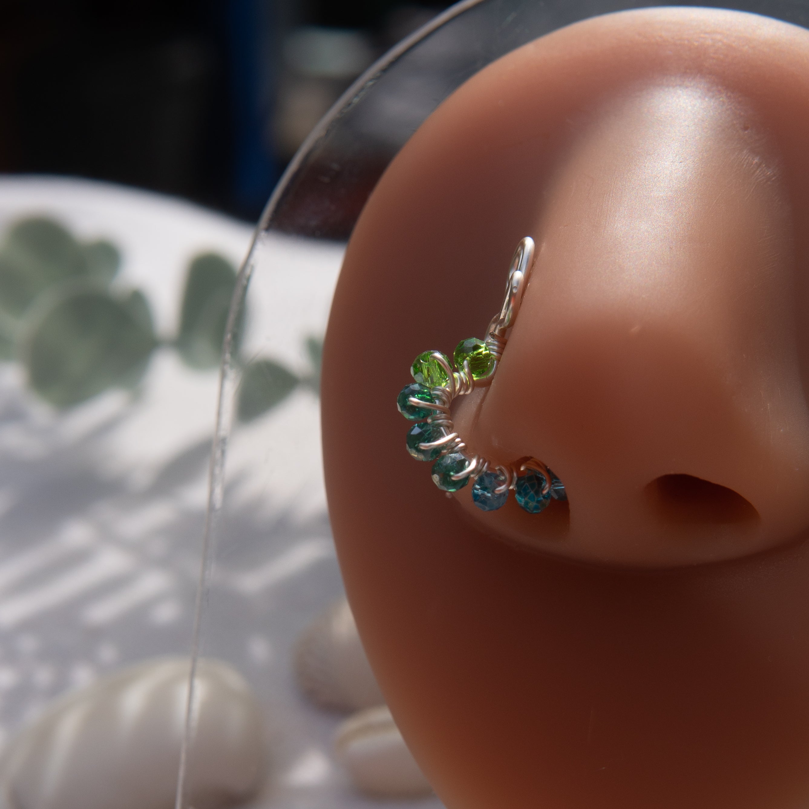 A silver-toned faux nose ring cuffs the model's nose. Transparent seed beads are wire-wrapped around the hoop's circumference. Above the hoop, the wire design forms an Ankh symbol.