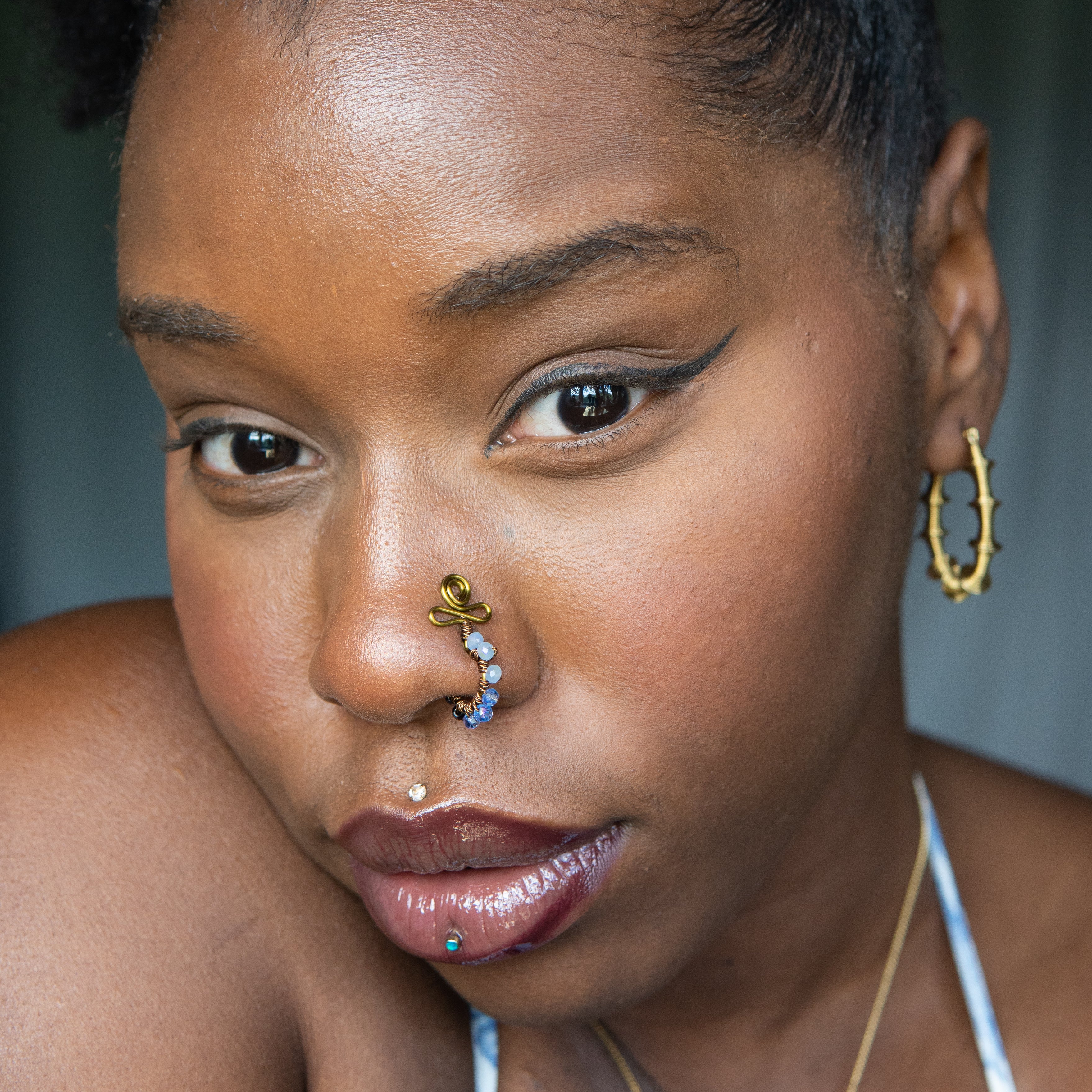 A bronze-toned faux nose ring cuffs the model's nose. Blue transparent seed beads are wire-wrapped around the hoop's circumference. Above the hoop, the wire design forms an Ankh symbol