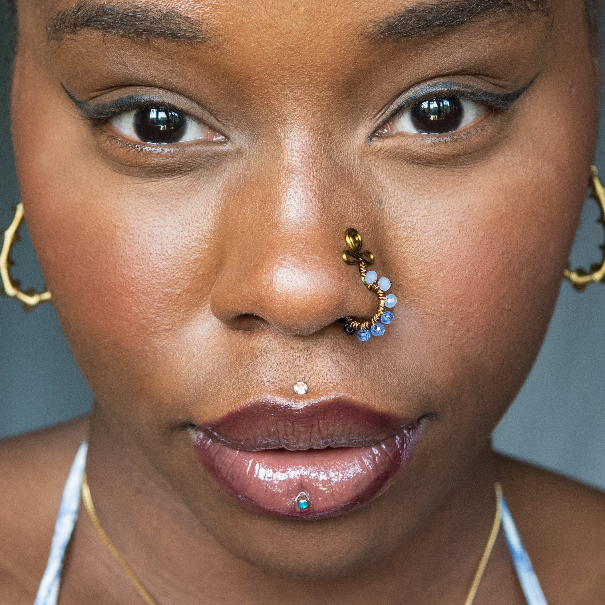 A bronze-toned faux nose ring cuffs the model's nose. Blue transparent seed beads are wire-wrapped around the hoop's circumference. Above the hoop, the wire design forms an Ankh symbol