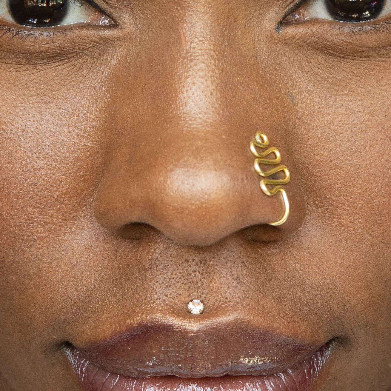 A gold-toned faux nose ring cuffs the model's nose. The wire design moulds a wavy pyramid shape.