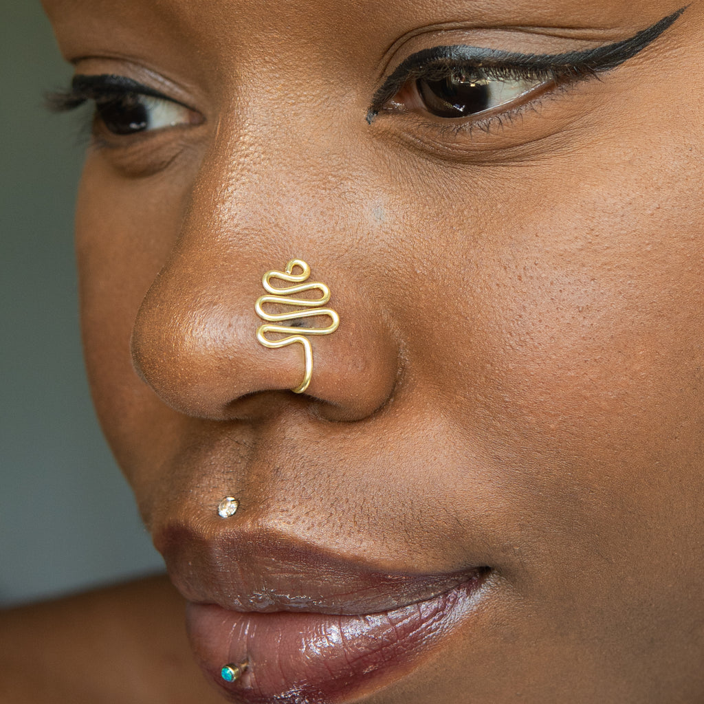A gold-toned faux nose ring cuffs the model's nose. The wire design moulds a wavy pyramid shape.