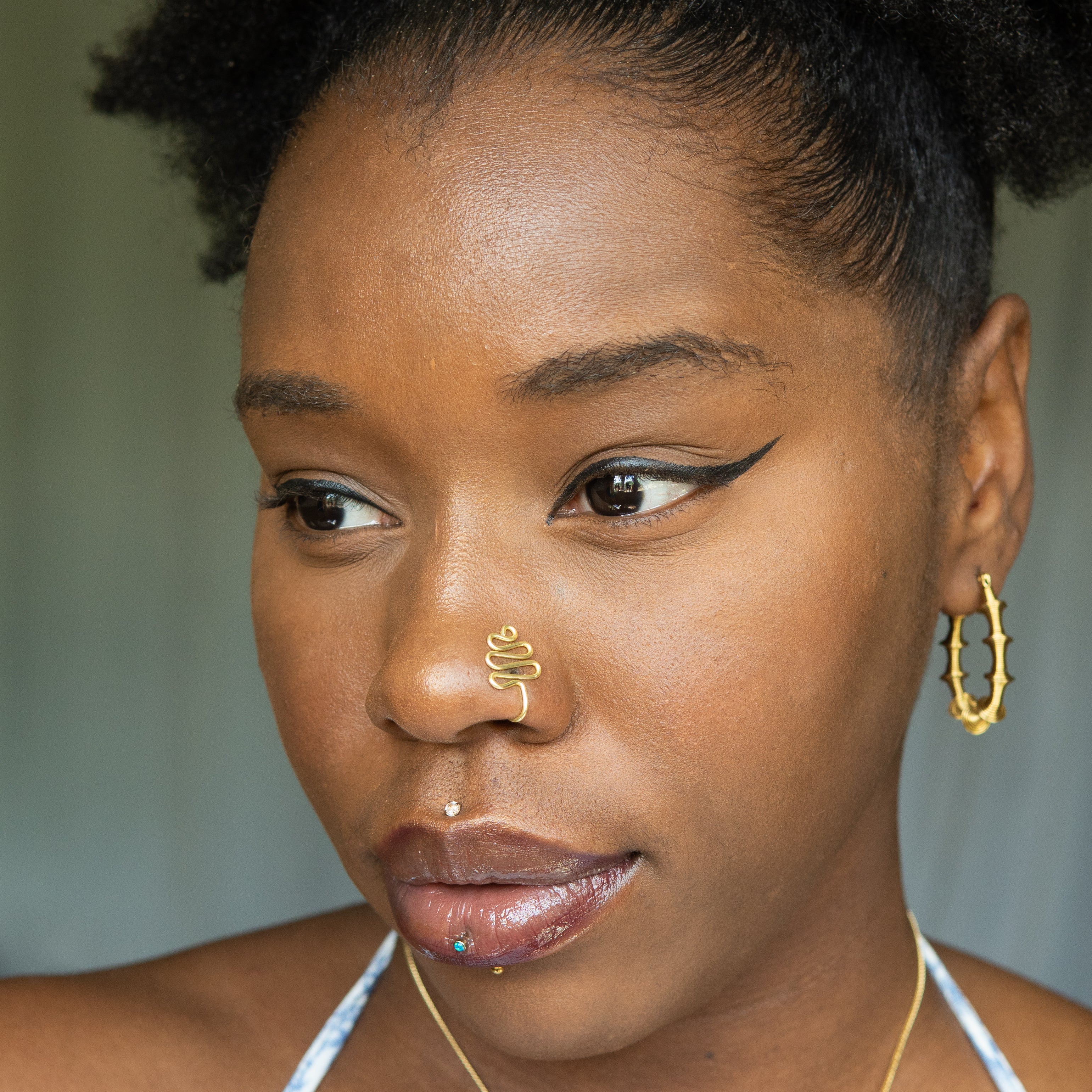 A gold-toned faux nose ring cuffs the model's nose. The wire design moulds a wavy pyramid shape.