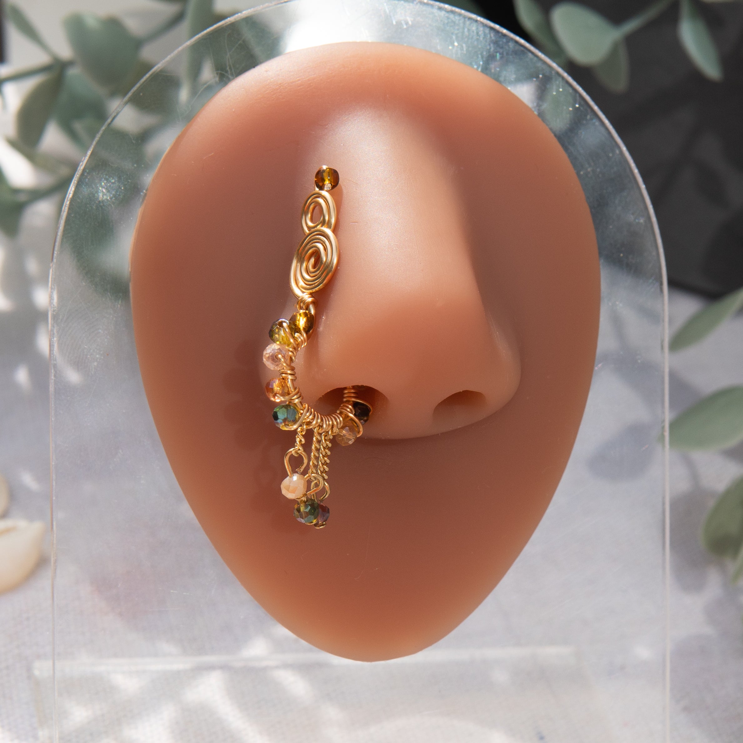 A gold-toned faux dangle nose ring cuffs the model's nostrils. Transparent mix of brown and green seed beads are wire-wrapped around the hoop's circumference, with three beaded gold curb chains dangling down.