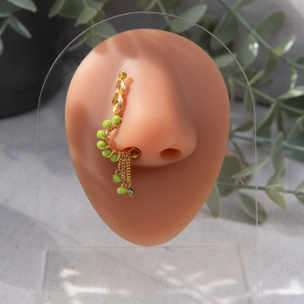 A light gold-toned faux dangle nose ring cuffs the model's nostrils. Green seed beads are wire-wrapped around the hoop's circumference, with three beaded gold chains dangling down.