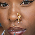 A gold-toned, non-pierced nose ring cuffs on the model's nostril. The wire design spirals up the nose, creating a pyramid shape. Two 18K gold curb chains dangle from the nose cuff, with lilac seed beads attached.