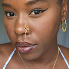 A gold-toned non-pierced nose ring cuffs on the model's nostril. The wire design spirals up the nose, creating a pyramid shape. Two 18K gold curb chains dangle from the nose cuff, with lilac seed beads attached.