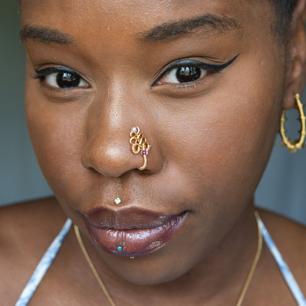 A gold-toned, non-pierced nose ring cuffs on the model's nostril. The wire design spirals up the nose, creating a pyramid shape with two seed beads (purple and lilac) weaved into the design.