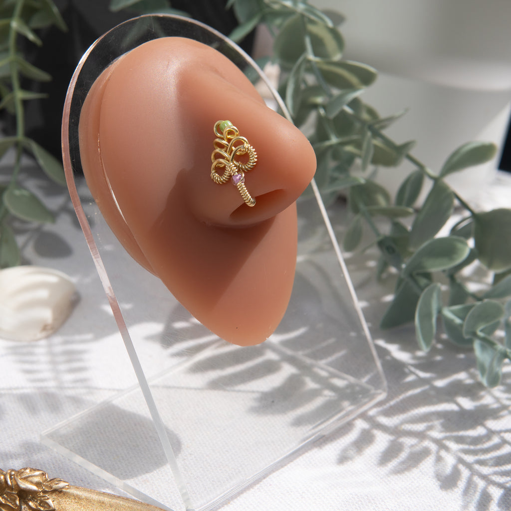 A light gold-toned, non-pierced nose ring cuffs on the model's nostril. The wire design spirals up the nose, creating a pyramid shape with two seed beads (green and lilac) weaved into the design.