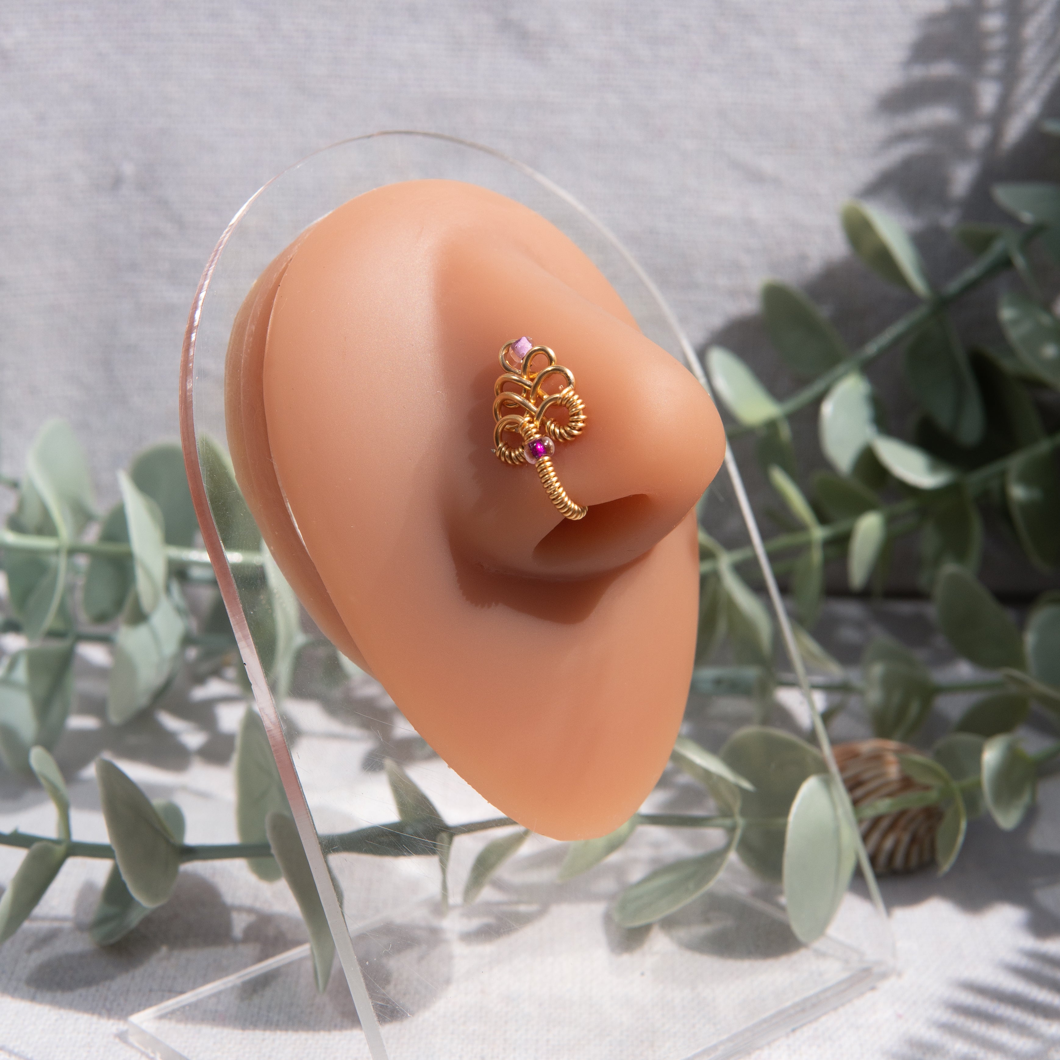 A gold-toned, non-pierced nose ring cuffs on the model's nostril. The wire design spirals up the nose, creating a pyramid shape with two seed beads (purple and lilac) weaved into the design.