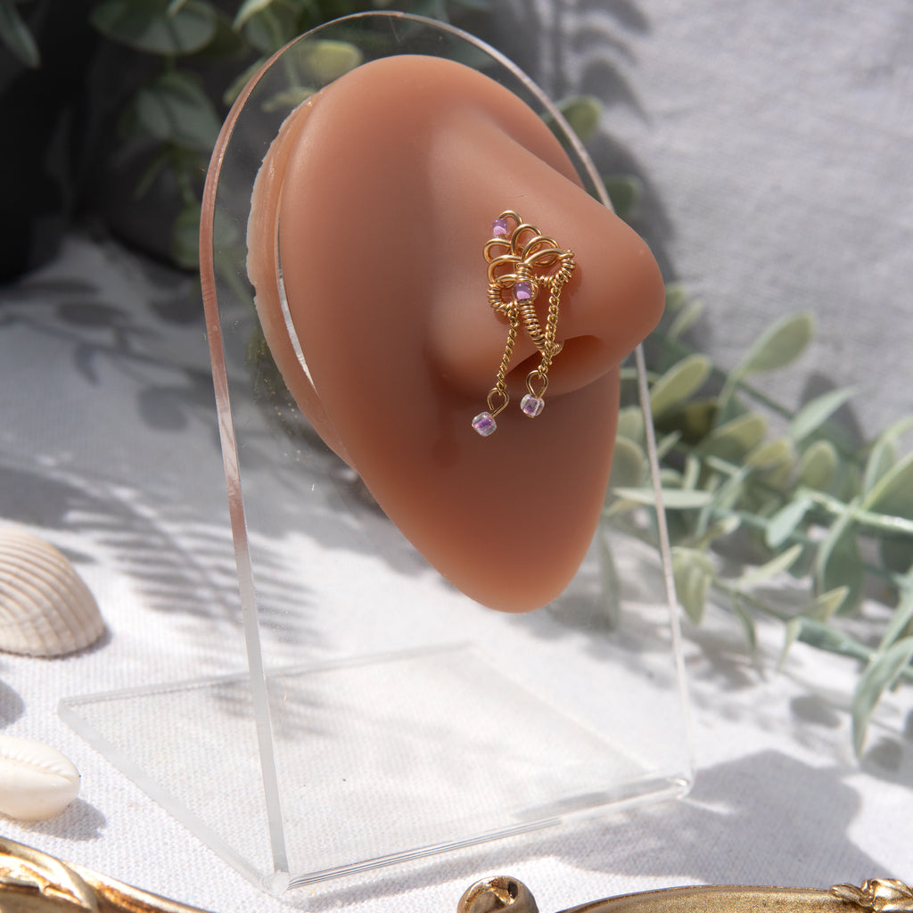 A gold-toned, non-pierced nose ring cuffs on the model's nostril. The wire design spirals up the nose, creating a pyramid shape. Two 18K gold curb chains dangle from the nose cuff, with lilac seed beads attached.