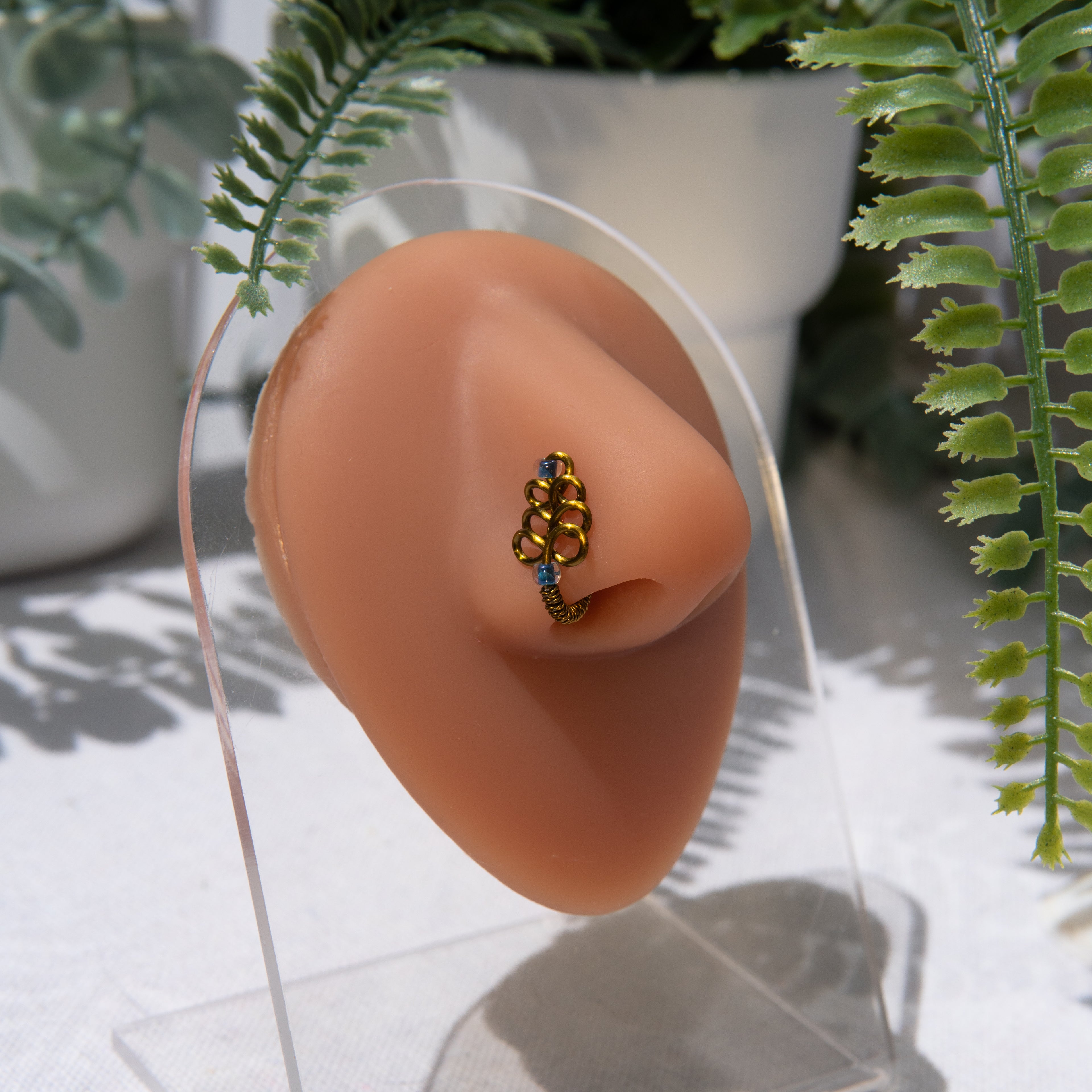 A bronze-toned, non-pierced nose ring cuffs on the model's nostril. The wire design spirals up the nose, creating a pyramid shape with two seed beads (blue) weaved into the design.