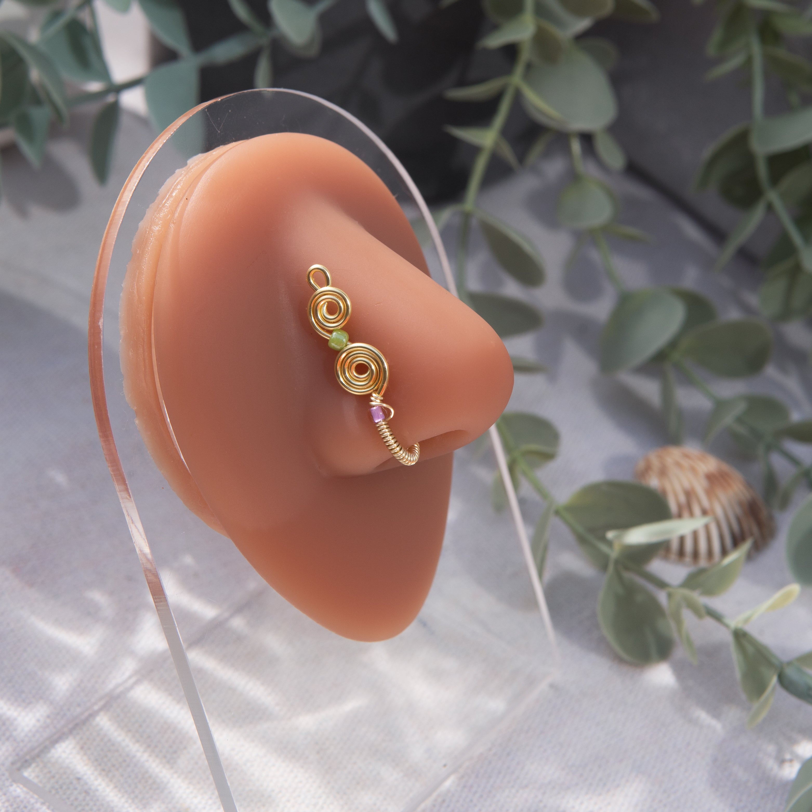 A light gold-toned, tightly spiralled nose cuff rests on the model's nose. Two seed beads (green and lilac) are interwoven into the design as the 3-tier circular spiral gradually decreases in size.
