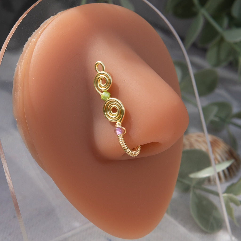 A light gold-toned, tightly spiralled nose cuff rests on the model's nose. Two seed beads (green and lilac) are interwoven into the design as the 3-tier circular spiral gradually decreases in size.