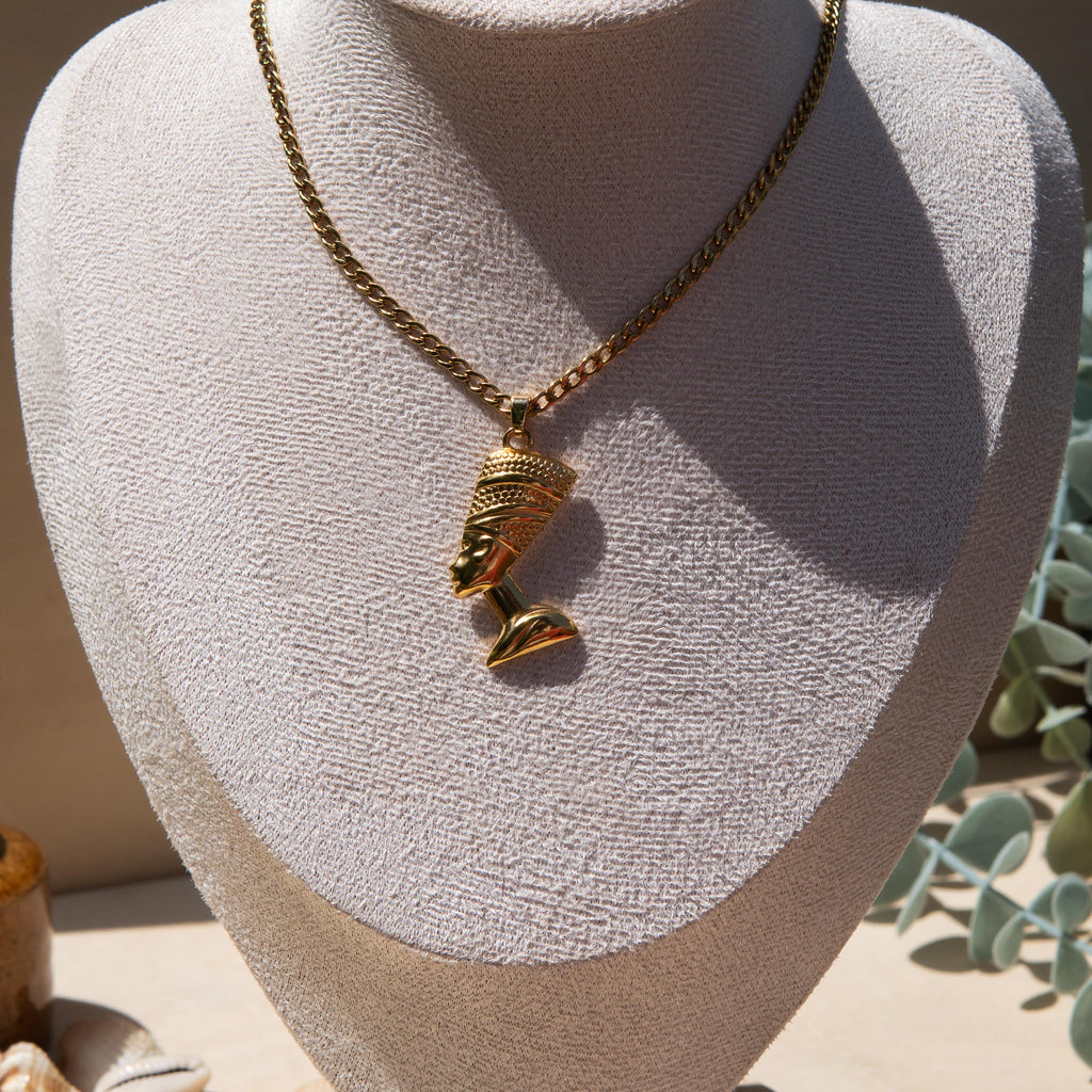 A gold-toned pendant necklace displayed on a textured off-white jewellery bust. The pendant is an Egyptian Queen Nefertiti