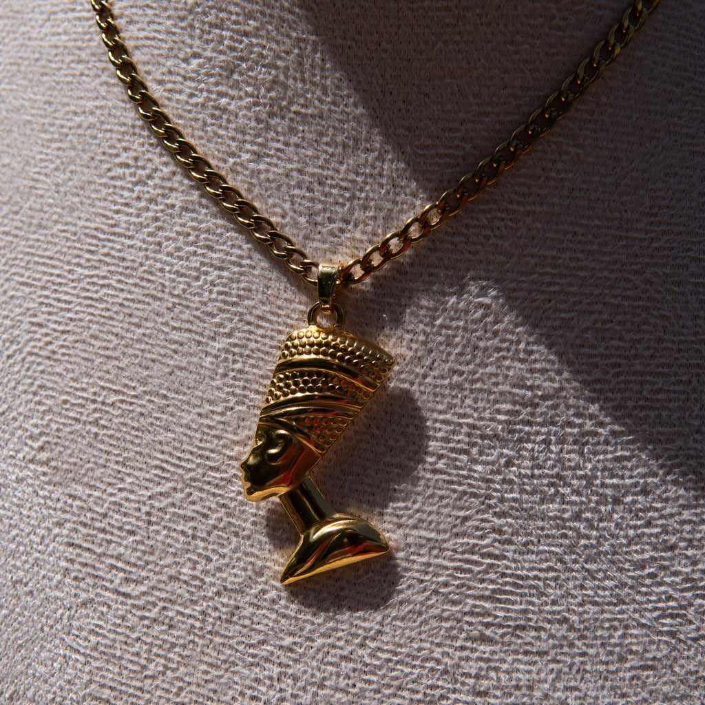 A gold-toned chain necklace with a Nefertiti head pendant, displayed on a textured light gray surface with strong sunlight and shadows