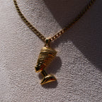 A gold-toned chain necklace with a Nefertiti head pendant, displayed on a textured light gray surface with strong sunlight and shadows