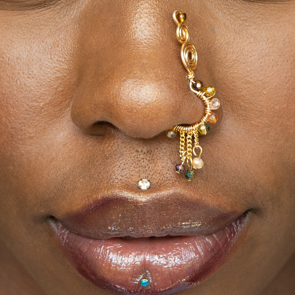 A gold-toned faux dangle nose ring cuffs the model's nostrils. A mix of brown and green transparent seed beads are wire-wrapped around the hoop's circumference, with three beaded gold curb chains dangling down.