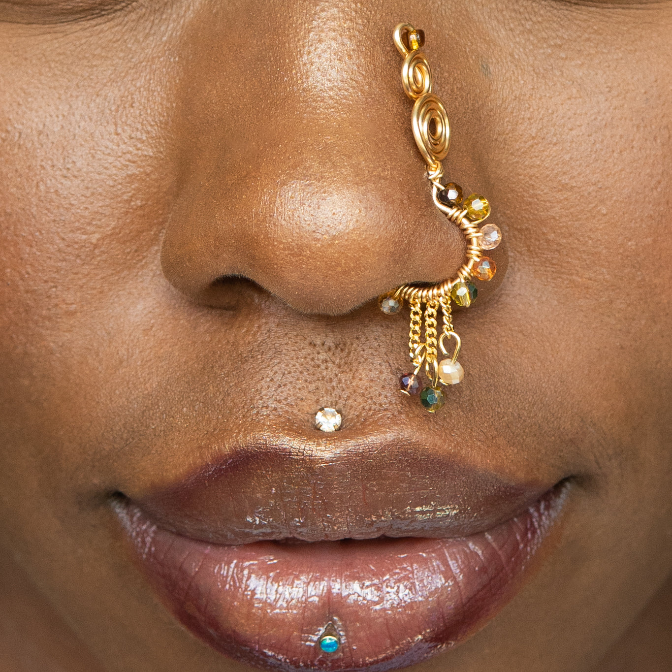 A gold-toned faux dangle nose ring cuffs the model's nostrils. A mix of brown and green transparent seed beads are wire-wrapped around the hoop's circumference, with three beaded gold curb chains dangling down.