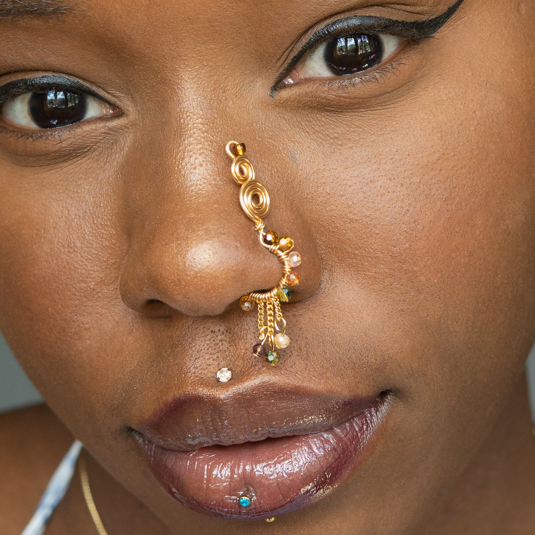 A gold-toned faux dangle nose ring cuffs the model's nostrils. A mix of brown and green transparent seed beads are wire-wrapped around the hoop's circumference, with three beaded gold curb chains dangling down.