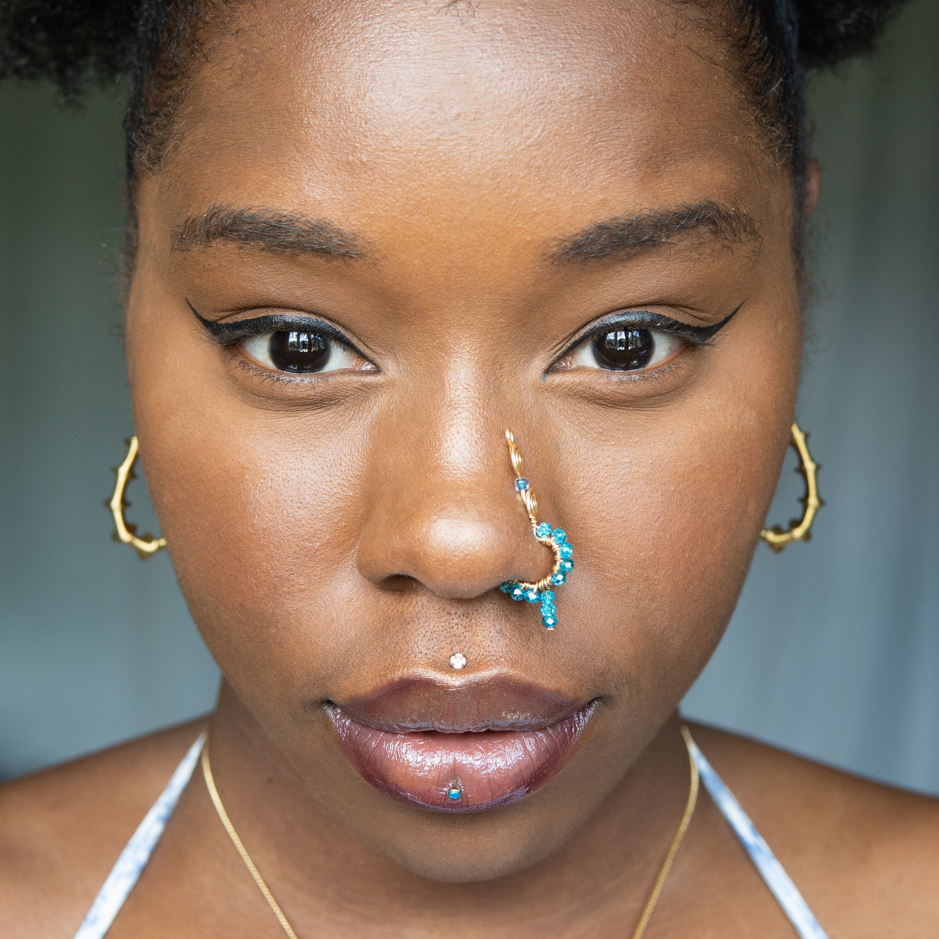A gold-toned faux dangle nose ring cuffs a model's nose. Blue transparent seed beads are wire-wrapped around the hoop's circumference, with a beaded wire dangling down.