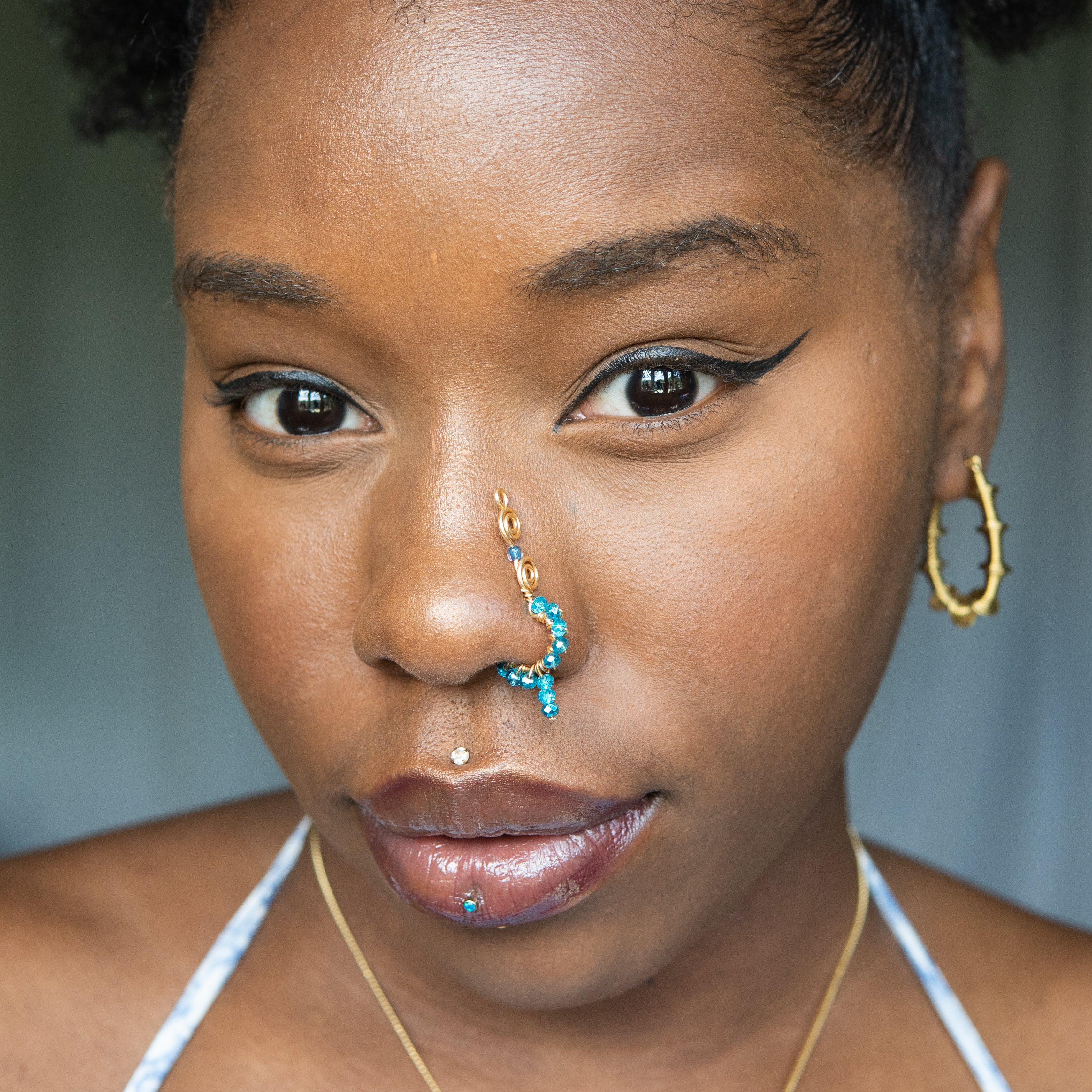 A gold-toned faux dangle nose ring cuffs a model's nose. Blue transparent seed beads are wire-wrapped around the hoop's circumference, with a beaded wire dangling down.