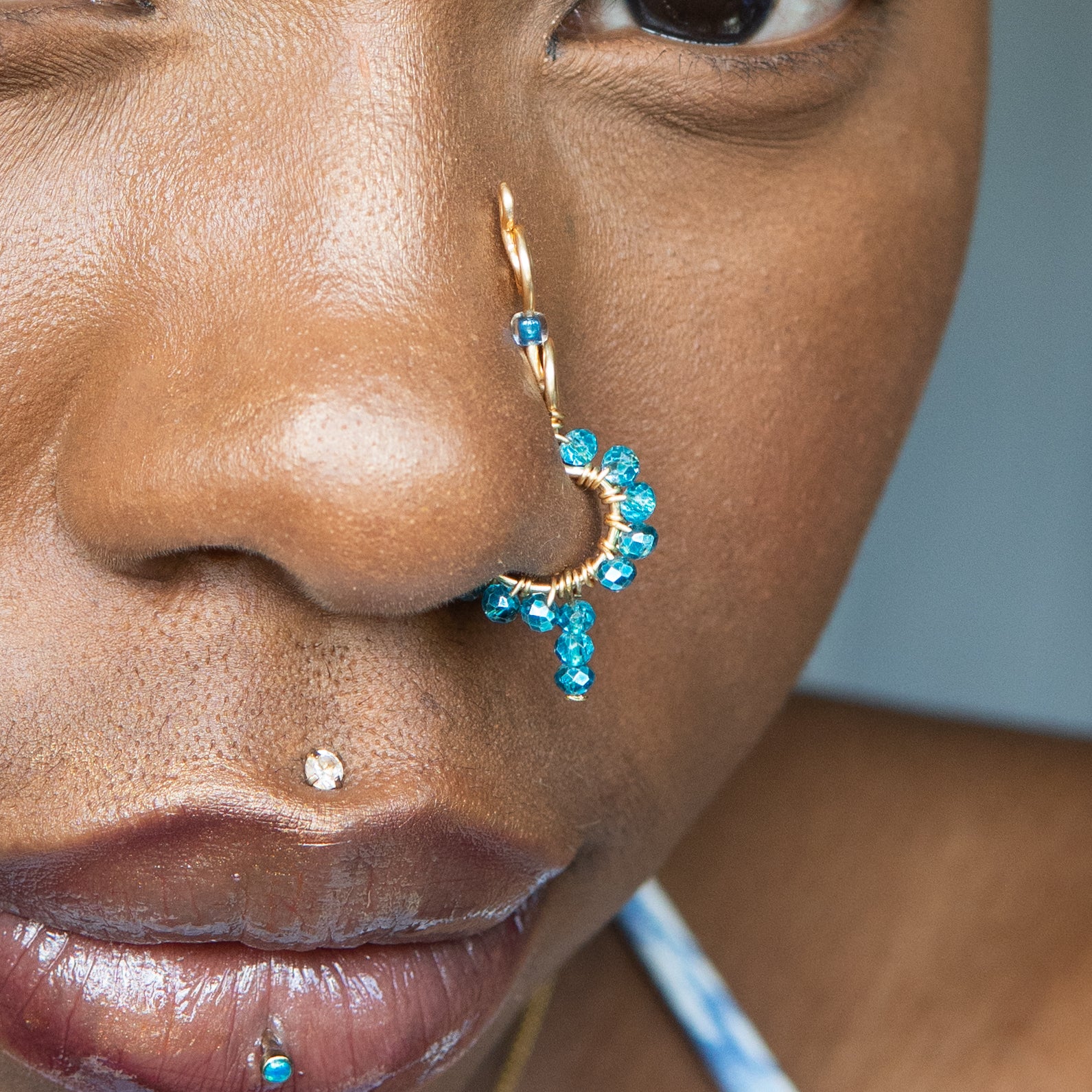 A gold-toned faux dangle nose ring cuffs a model's nose. Blue transparent seed beads are wire-wrapped around the hoop's circumference, with a beaded wire dangling down.
