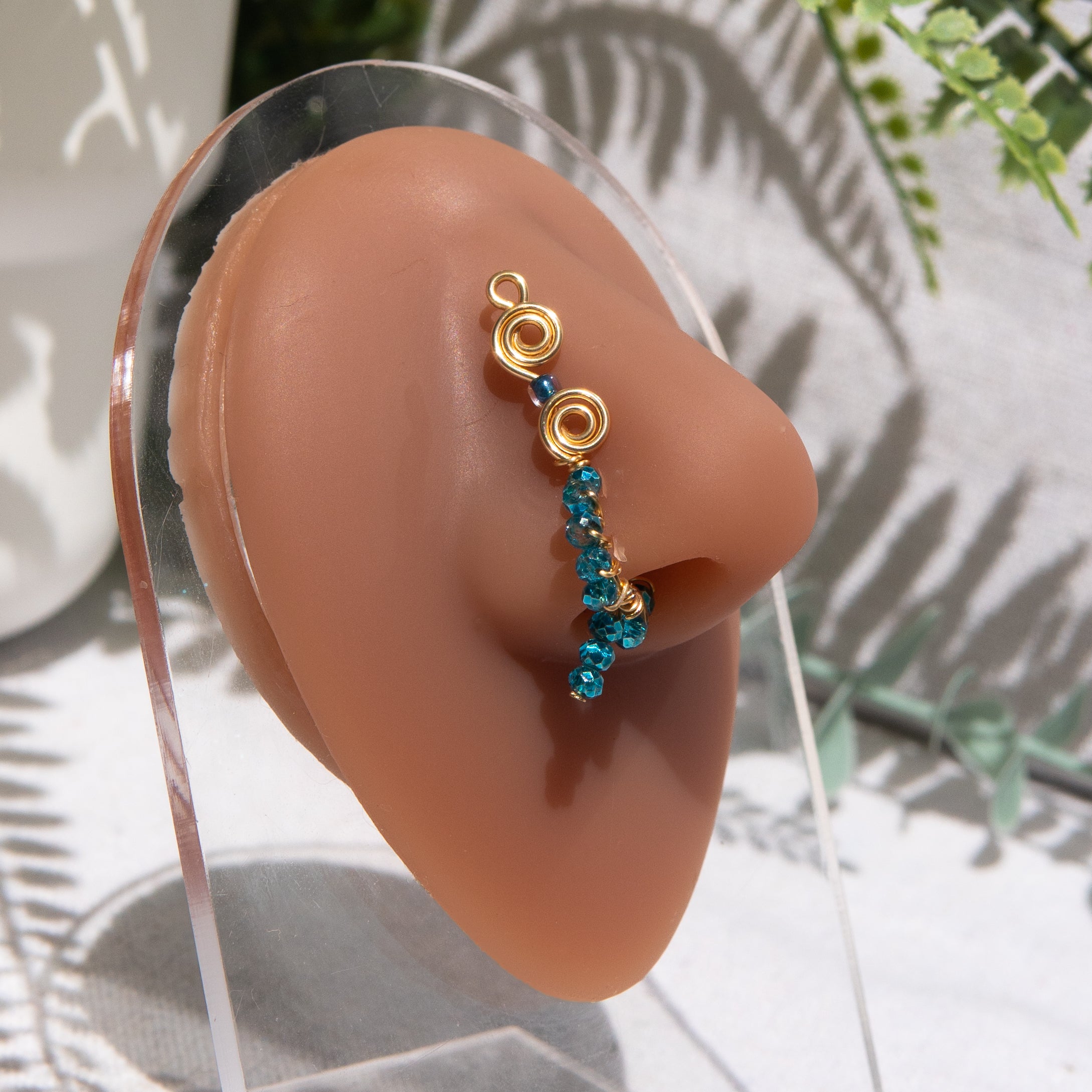 A gold-toned faux dangle nose ring is cuffed on a model's nose. Blue transparent seed beads are wire-wrapped around the hoop's circumference, with a beaded wire dangling down.