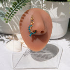 A gold-toned faux dangle nose ring is cuffed on a model's nose. Blue transparent seed beads are wire-wrapped around the hoop's circumference, with a beaded wire dangling down.