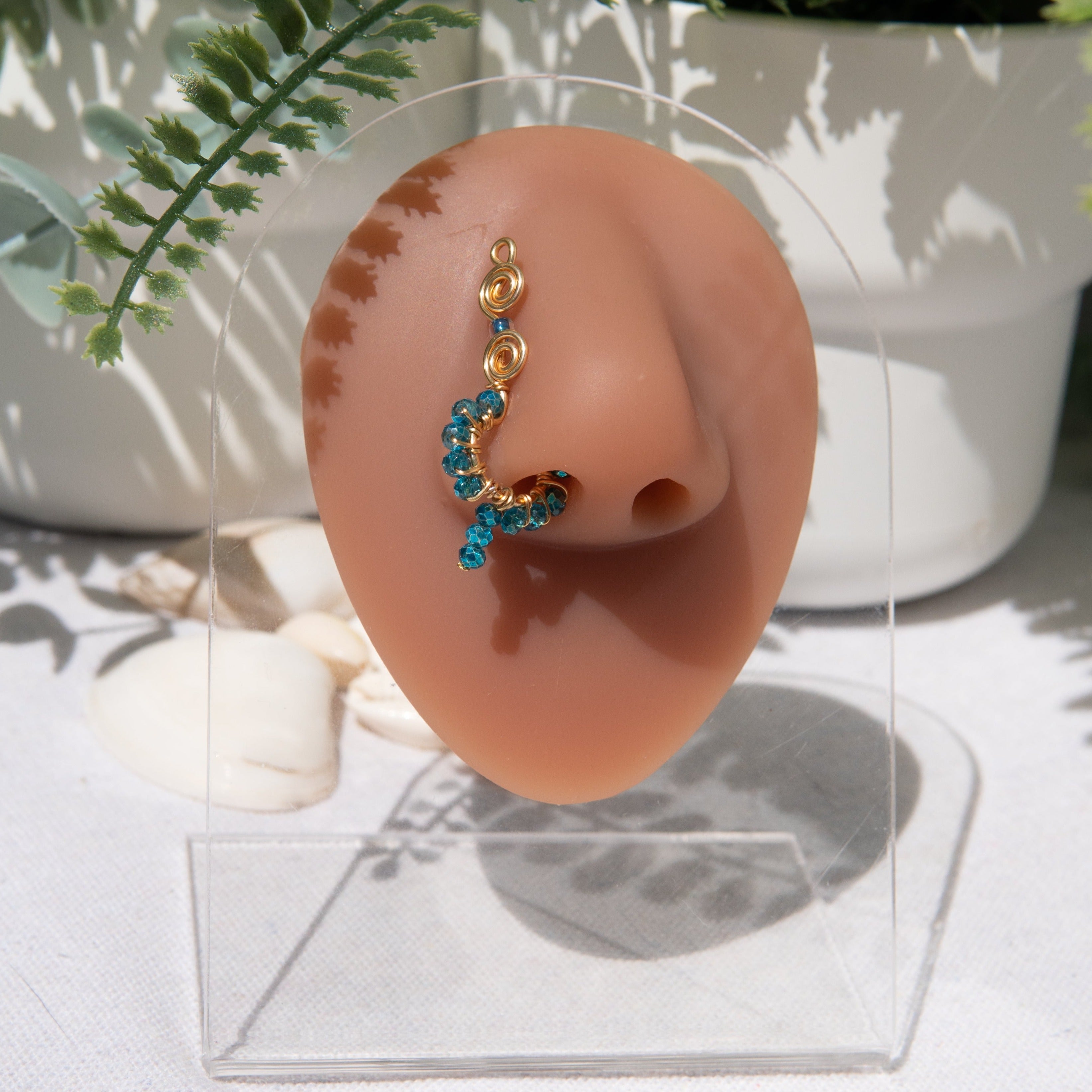 A gold-toned faux dangle nose ring is cuffed on a model's nose. Blue transparent seed beads are wire-wrapped around the hoop's circumference, with a beaded wire dangling down.