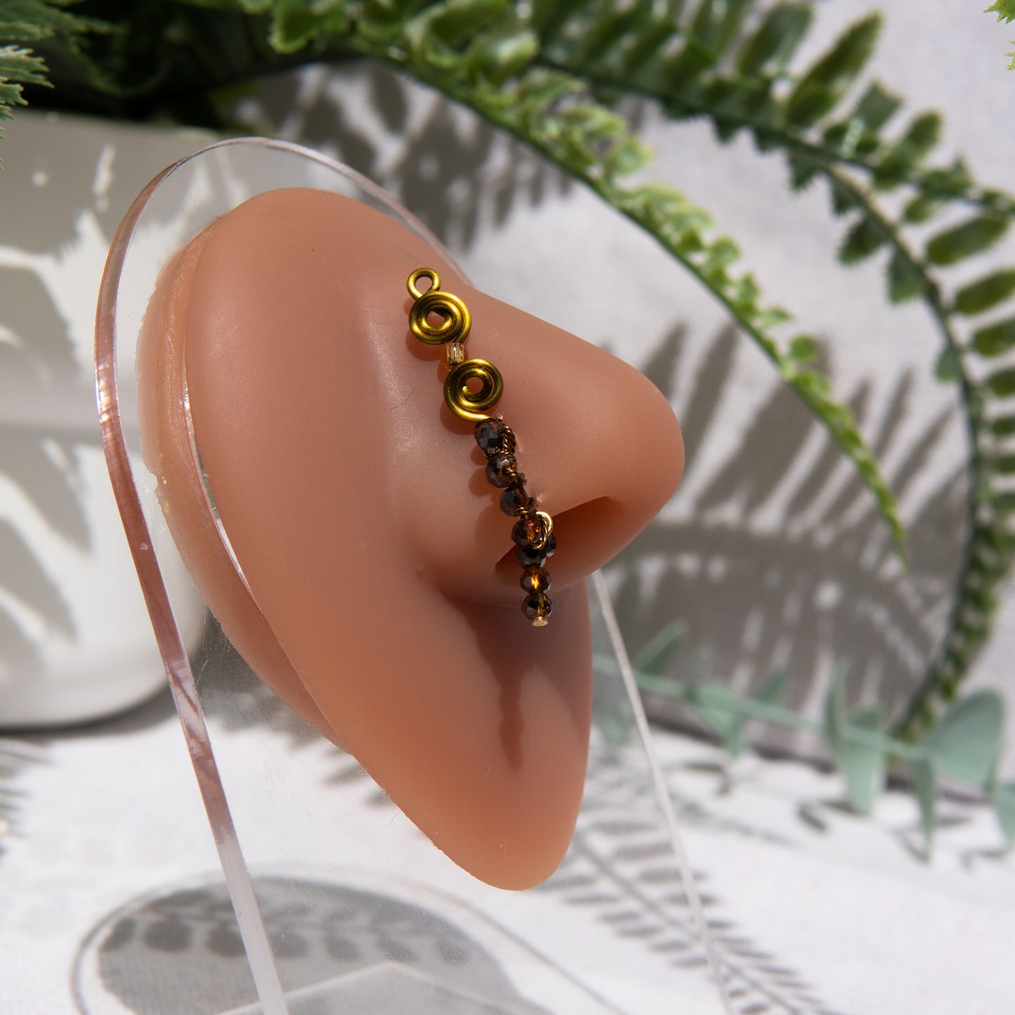 A bronze-toned faux dangle nose ring cuffs a model's nose. Brown transparent seed beads are wire-wrapped around the hoop's circumference, with a beaded wire dangling down.